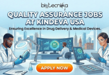 Quality Assurance Jobs in USA | Explore the Latest Job Openings at Kindeva Quality Assurance Jobs in USA. A high-quality recruitment banner for Biotecnika Global titled "QUALITY ASSURANCE JOBS AT KINDEVA USA" in bold white text. The subtitle reads "Ensuring Excellence in Drug Delivery & Medical Devices." The illustration shows a diverse team of scientists in a clean, modern laboratory. One researcher inspects an inhaler device with a magnifying glass, another monitors a large curved screen displaying "QC APPROVED," and a third professional tracks data on a tablet. Large windows in the background overlook a modern USA city skyline. A prominent orange "APPLY NOW" button is at the bottom.