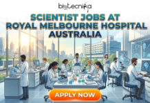 Scientist Jobs at Royal Melbourne Hospital Australia | Job You Can’t Miss in 2026 SCIENTIST JOBS AT ROYAL MELBOURNE HOSPITAL AUSTRALIA. A recruitment banner for Biotecnika Global, featuring the title "SCIENTIST JOBS AT ROYAL MELBOURNE HOSPITAL AUSTRALIA" in blue and gold text. The illustration depicts a team of diverse scientists working in a modern laboratory with a view of a Melbourne cityscape, including a hospital and a tram, through large windows. An "APPLY NOW" button is located at the bottom.