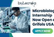 Microbiology Internship Now Open at Grifols USA | Future Microbiologists Wanted! Microbiology Internship Now Open at Grifols USA. An advertisement for a "Microbiology Internship Now Open at Grifols USA" by Biotecnika Global. The top left features the Biotecnika Global logo. Below it is an image of a male scientist in a lab coat and safety glasses looking into a microscope, with Petri dishes and glassware on the bench. On the right, large text announces the internship, with a green and blue gradient "APPLY NOW" button below it.