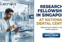 Research Fellowship in Singapore at National Dental Centre – Apply Now Research Fellowship in Singapore at National Dental Centre – Apply Now. A professional promotional image for a Research Fellowship in Singapore at National Dental Centre. On the left side, a male researcher in a white lab coat is shown in a high-tech dental laboratory, holding a 3D dental model while looking at a tablet. The background features a modern clinic setting with a large window revealing the iconic Singapore skyline, including the Marina Bay Sands. The right side of the graphic has a clean white background with bold, professional typography. At the top left is the Biotecnika Global logo. The main text reads "RESEARCH FELLOWSHIP IN SINGAPORE AT NATIONAL DENTAL CENTRE" in dark blue and gold, followed by the tagline "Advance your career in oral health research." At the bottom right, there is a prominent, glowing blue "APPLY NOW" button.