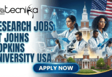 High-Paying Research Jobs in USA | Johns Hopkins University Hiring Now Research Jobs at Johns Hopkins University, USA. A featured image for a job advertisement titled "RESEARCH JOBS AT JOHNS HOPKINS UNIVERSITY USA." The graphic features a team of diverse scientists in white lab coats conducting high-level medical research in a state-of-the-art laboratory. They are using advanced equipment and interacting with glowing holographic displays of the human anatomy and viral structures. In the background, the iconic Gilman Hall and the Johns Hopkins University campus are depicted alongside a large American flag. The Biotecnika Global logo is prominently placed at the top left, and a bright blue "Apply Now" button is at the bottom center.