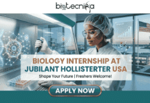 Biology Internship in USA: Earn $25/hr at Jubilant – Limited Seats! Biology Internship in USA Earn $25hr at Jubilant – Limited Seats! A promotional banner advertising a "BIOLOGY INTERNSHIP AT JUBILANT HOLLISTERSTIER USA." The image features a female scientist of color in full lab safety gear (hairnet, goggles, gloves, lab coat) in a modern laboratory, pipetting liquid into a multi-well plate within a laminar flow hood. To her left is a large, glowing green and blue hologram of a DNA double helix and complex molecular structures. Multiple augmented reality screens are visible; one on the right shows a positive-trend "Cell growth" chart and molecular data (with text like "C6H10"). Large windows in the background overlook a view of New York City, featuring the Statue of Liberty, an American flag, and a modern building. Central text below the title reads, "Shape Your Future | Freshers Welcome!". A prominent "APPLY NOW" button is at the bottom center. A footer text reads, "Launch Your Scientific Journey in the USA | Global Opportunities." The Biotecnika logo is at the top center.
