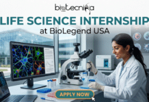Life Science Internship in USA – Join BioLegend as a Product Development Intern! Life Science Internship at BioLegend USA | Product Development. A promotional banner with the biotecnika Global logo at the top center. Below the logo, large text reads 'LIFE SCIENCE INTERNSHIP' with 'at BioLegend USA' below that. The main scene depicts a young woman in a lab coat and blue gloves, smiling as she works with a micropipette to dispense liquid into a multi-well plate. She is seated at a desk cluttered with laboratory equipment, including a microscope and numerous test tubes. Behind her, a large computer monitor displays a vibrant, colorful fluorescent microscope image of a biological sample, showing intricate, branching structures with colors like blue, green, and red. In the background, through a large window, a modern biotech building is visible under a clear sky, with a world map pattern overlaid on the window's view. To the right of the desk, a specimen wall features various biological samples with scientific labels. At the bottom center, a prominent orange and teal button reads 'APPLY NOW' and has a mouse cursor clicking on it. The overall setting is professional, inviting, and high-tech.