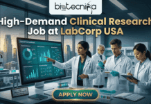 High-Demand Clinical Research Job at LabCorp USA for Life Science Graduates! High-Demand Clinical Research Job at LabCorp USA for Life Science Graduates!. A promotional featured image from biotecnika Global with the headline 'High-Demand Clinical Research Job at LabCorp USA'. The image depicts a modern, high-tech laboratory with clean lines and high windows overlooking a large city skyline at dusk. A diverse team of four clinical research scientists (one woman in the foreground and three men in the background) in white lab coats are working. The central female scientist points to a large computer monitor displaying a detailed 'Clinical Trial Dashboard' with various charts, graphs, and molecular pathway diagrams. Below the screen, a plaque on the desk reads 'USA Clinical Research Excellence'. Other scientists are observing, pipetting into a well plate, and taking notes on a clipboard. At the bottom center, a prominent button with golden trim reads 'APPLY NOW'.