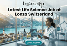 Latest Life Science Job in Switzerland | Lonza Hiring PhD Candidates Latest Life Science Job in Switzerland Lonza Hiring PhD Candidates. A promotional image for a job posting from Biotecnika Global, featuring the large text headline 'Latest Life Science Job at Lonza Switzerland' in the upper center. Below the headline is a scene of a modern laboratory with a female scientist of color wearing safety glasses and a lab coat. She is holding a tablet that displays a DNA helix. She looks from the data towards a very large window that overlooks a picturesque Swiss mountain landscape with a lake and a small town. In the foreground is a lab workstation with a glass bioreactor and complex chemical formulas. To the right, a robot arm and high-tech monitors are visible. A prominent teal button at the bottom center reads 'APPLY NOW'."