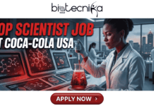 Scientist Job Alert: Coca-Cola Hiring Product Development Scientist – Top Food Science Jobs in USA Scientist Job Alert Coca-Cola Hiring Product Development Scientist – Top Food Science Jobs in USA. A professional featured image banner for a "Top Scientist Job at Coca-Cola USA". The biotecnika Global logo is at the top center. The main visual is an attractive photo of a Black female scientist in a white lab coat and blue gloves, focused on carefully pipetting a dark, amber-colored bubbling liquid into a heated stir-plate flask on a high-tech console in a modern laboratory. Multiple glowing screens on the console display detailed red molecular structures and charts related to formulation and carbonation. Other scientists are visible in the slightly blurred background, which features a large window overlooking a city skyline at dusk. A prominent, glossy red button at the bottom center clearly states "APPLY NOW >".