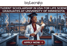 Life Science Student Scholarship at University of Minnesota USA | Apply Before Deadline Student Scholarship in USA for Life Science Graduates Apply Before Deadline. An illustrative image from biotecnika Global titled "STUDENT SCHOLARSHIP IN USA FOR LIFE SCIENCE GRADUATES AT UNIVERSITY OF MINNESOTA." The scene depicts a diverse female graduate student in a lab coat and graduation cap smiling as she holds a diploma which explicitly states: "LIFE SCIENCE SCHOLARSHIP AWARD - U OF MN." She stands in a sophisticated life sciences laboratory, surrounded by advanced research equipment like microscopes, computers displaying cell analysis, and molecular models. Through a large window behind her is a panoramic view of the University of Minnesota campus and the Minneapolis skyline across the Mississippi River at dusk. A prominent red "APPLY NOW →" button is located at the bottom center.