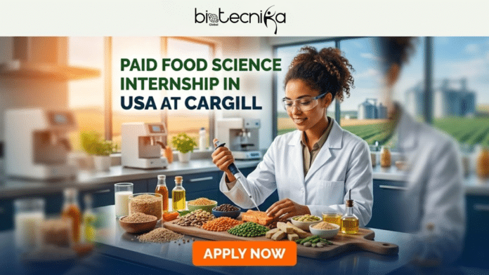 Paid Food Science Internship in USA at Cargill | Earn Upto $30/Hr. A featured job alert graphic from Biotecnika Global advertising a 'Paid Food Science Internship in USA at Cargill', with a woman in a white lab coat and safety glasses working with pipettes and a multi-plate display of whole food ingredients in a modern test kitchen setting. The background includes windows overlooking a farm, other kitchen equipment, and a blurred person. A large orange button at the bottom says 'APPLY NOW'.